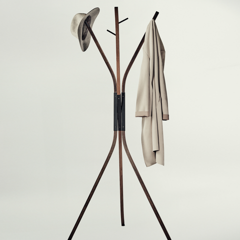 A modern wooden coat rack with three legs and curved top arms, shown against a plain light gray background. A hat is hanging on the left hook and a light beige coat is hanging on the right.