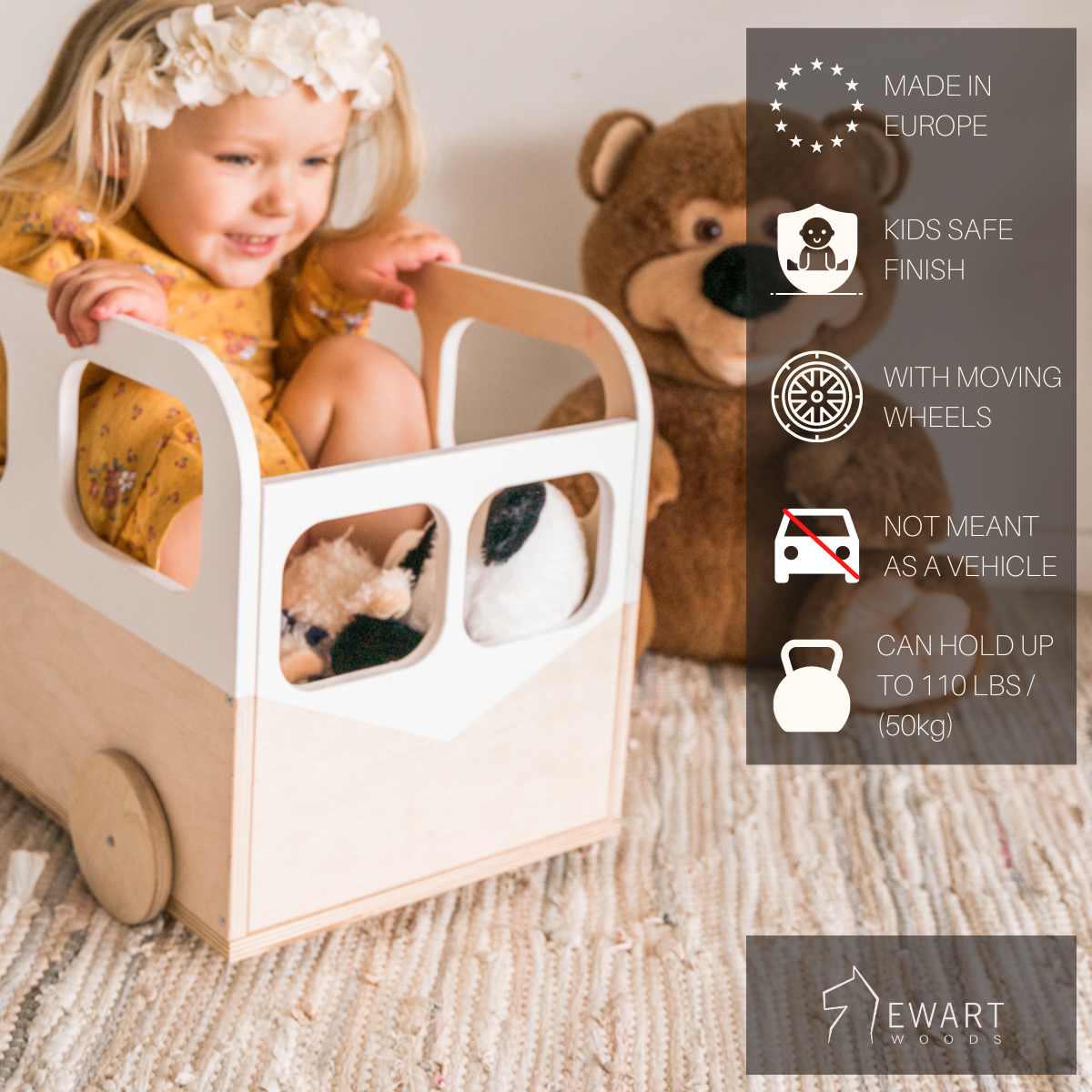 A young girl sitting inside a wooden toy bus with white accents and moving wheels, holding stuffed animals, with a teddy bear in the background on a cozy rug. The bus has a smooth, kid-safe finish and can hold up to 110 lbs.
