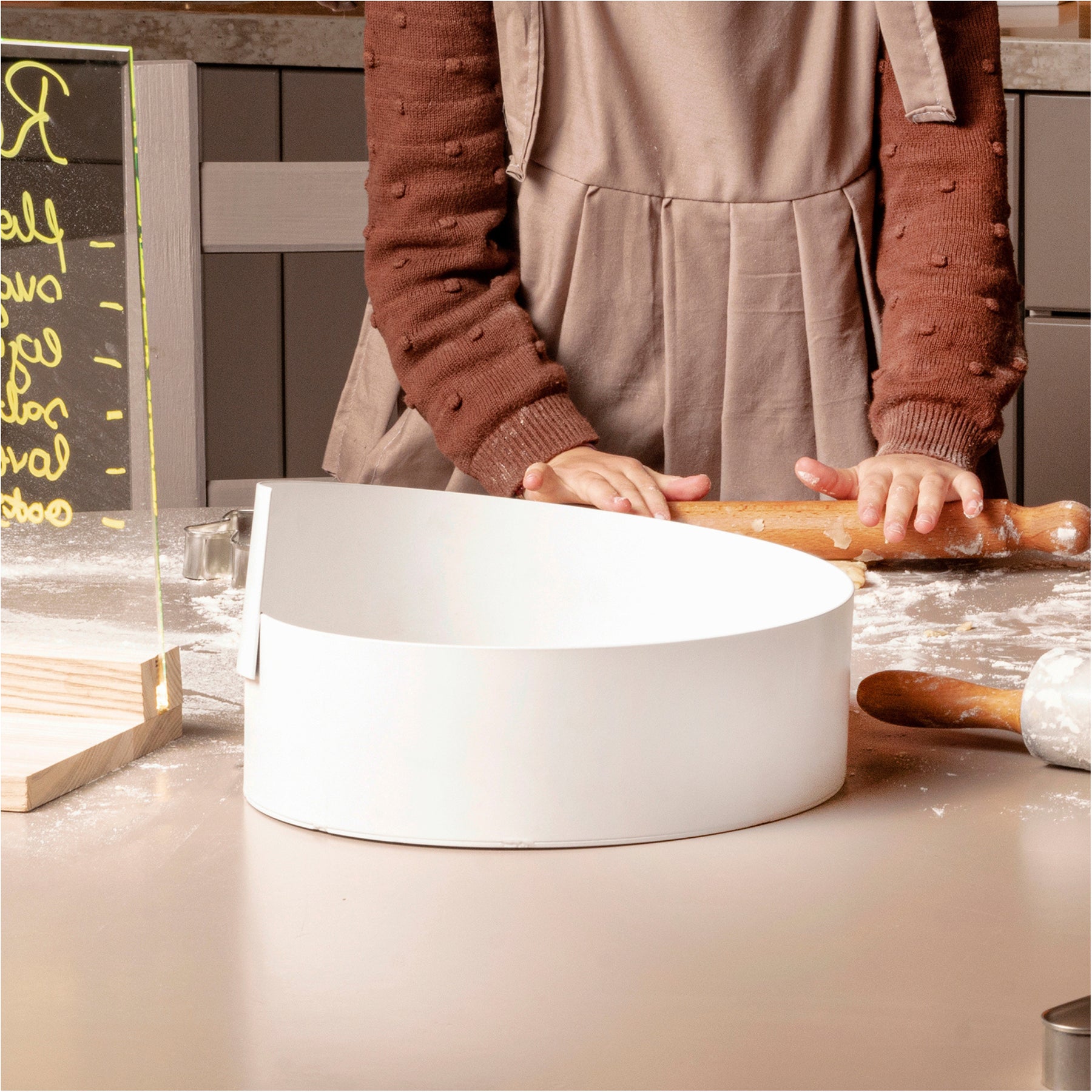 White metal bowl with a modern, minimalist design placed on a kitchen counter, surrounded by baking tools, while a person in an apron prepares dough.