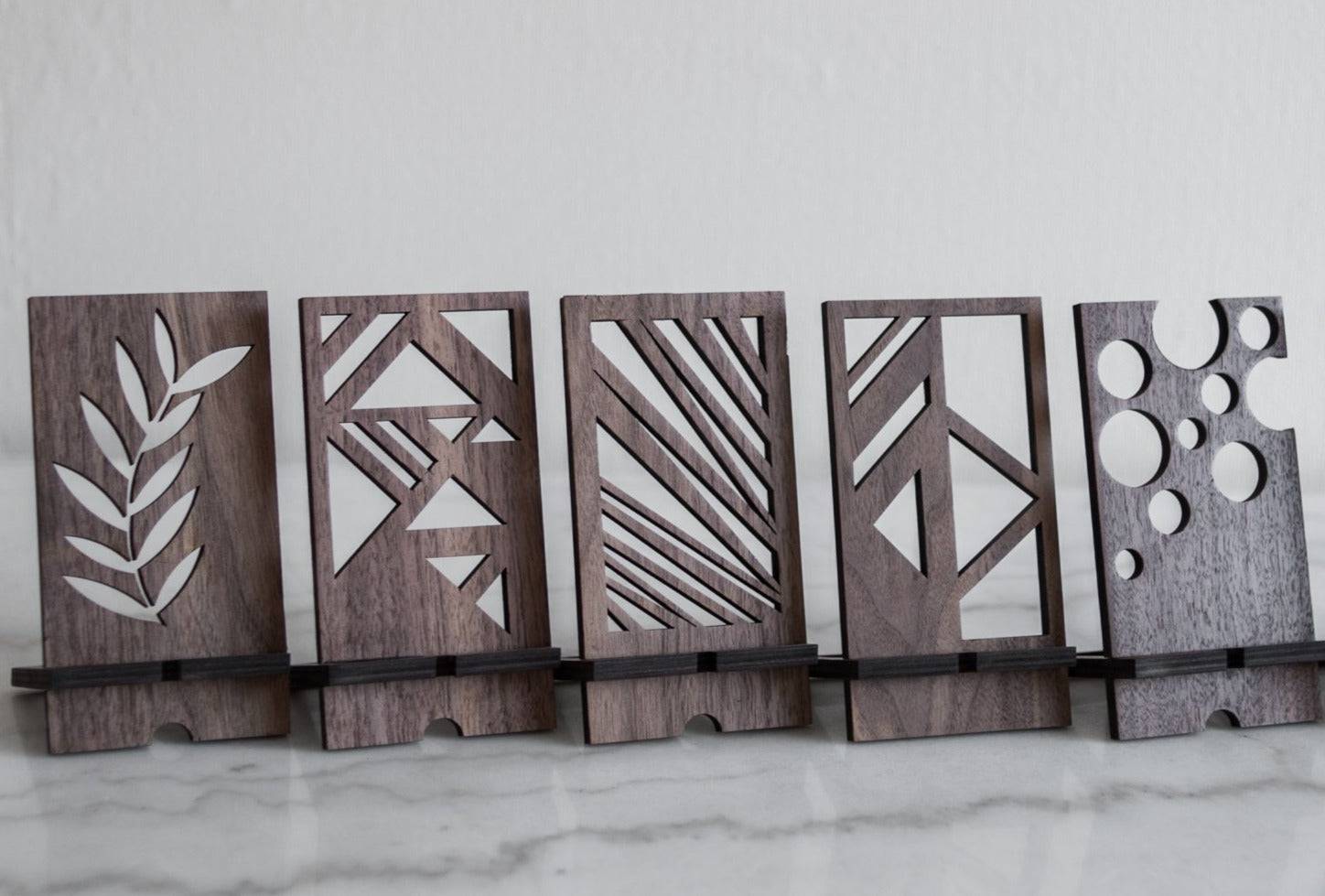 Wooden phone stands in walnut finish with unique geometric cutout designs: Leaf pattern, Abstract triangles, Diagonal lines, Geometric shapes, Circular cutouts. Each stand holds a phone at an angle on a smooth surface.