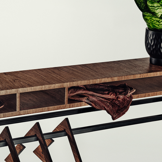 Close-up of a dark wood console table with open cubbies, a draped brown leather accessory, geometric wooden legs, and a potted plant on a light neutral background.