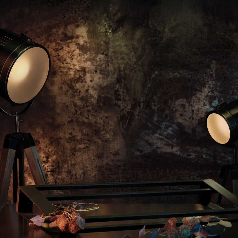 Industrial-style black floor lamps with warm white light on a dark rustic textured wall, with a black metal footwear holder in the foreground and dried flowers at the bottom.
