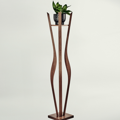A tall sculptural wooden plant stand with a rectangular base and two curved vertical supports, holding a small potted green plant on a narrow shelf. The stand appears to be made of medium-to-dark brown wood, photographed against a plain light gray studio background.
