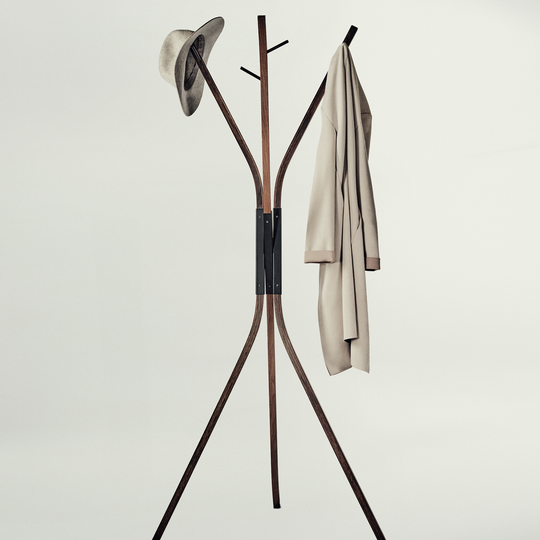 A modern wooden coat rack with three legs and curved top arms, shown against a plain light gray background. A hat is hanging on the left hook and a light beige coat is hanging on the right.