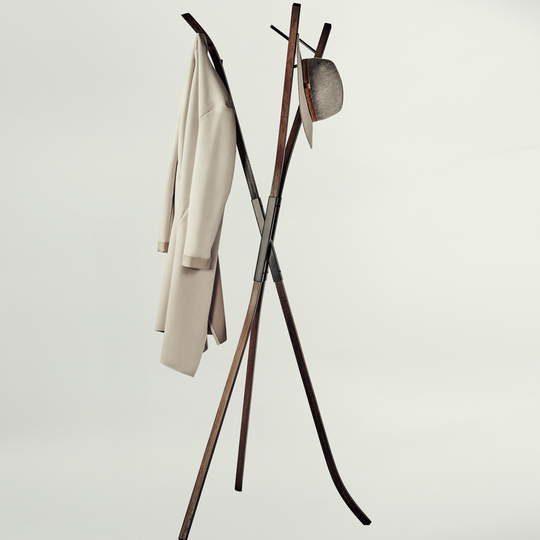 A modern wooden coat rack with an X-shaped base and curved top arms, shown against a plain light gray studio background. A light beige coat hangs on the left hook, and a textured gray hat is suspended on the right.