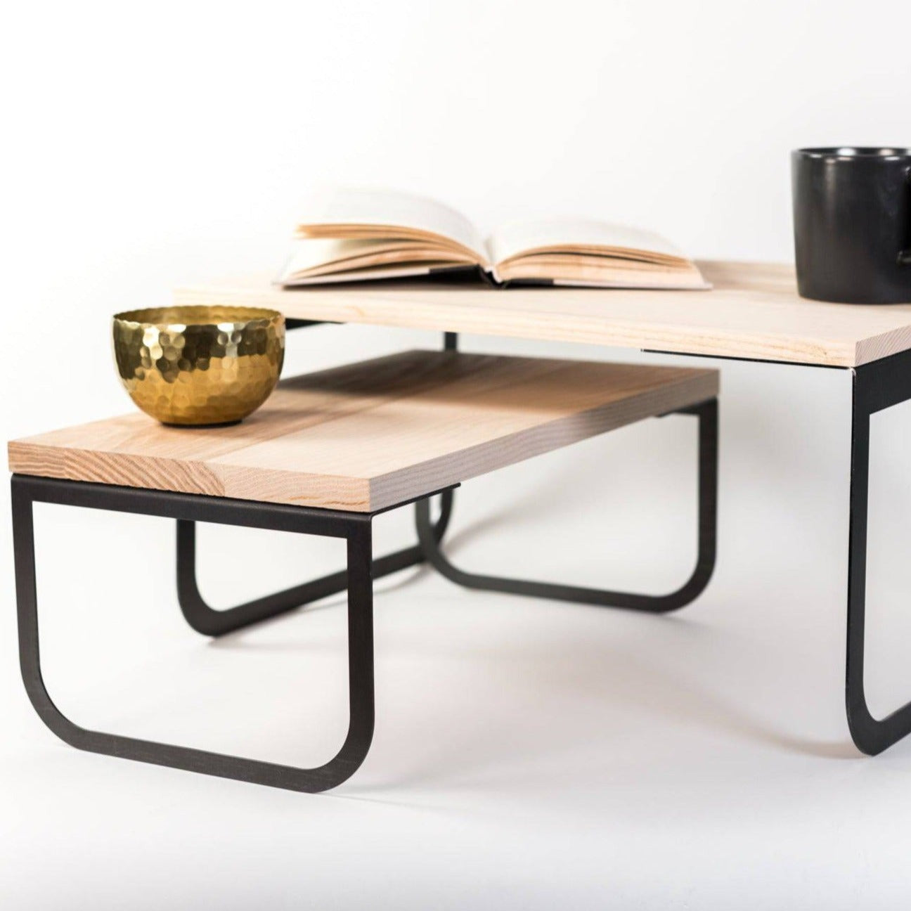 Modern wooden coffee table with black metal legs, featuring an open book, a gold bowl, and a black mug on its surface, isolated on a white background.