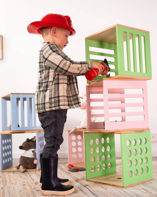 A child wearing a firefighter helmet and boots holds a toy fire extinguisher aimed at a stack of colorful wooden blocks with various cut-out designs, including slats and circular holes.